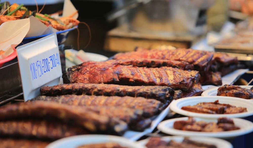 Juicy barbecue ribs with a glossy glaze displayed on a table at a Bangkok street food stall, with a variety of other local dishes in the background, inviting passersby to savor the rich and smoky flavors.