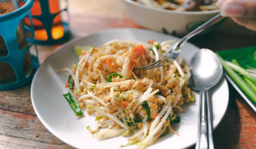A plate of freshly prepared Pad Thai with a tantalizing mix of noodles, bean sprouts, and shrimp, garnished with a lime wedge, ready to be enjoyed at a rustic wooden table, highlighting the simple yet rich flavors of Thai street cuisine.