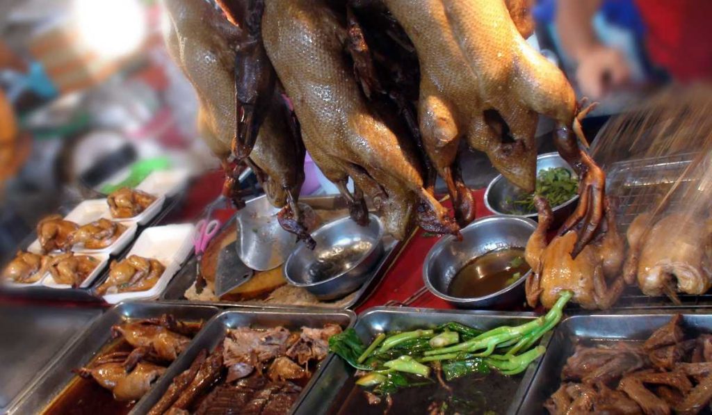 A close-up of a Bangkok street food stall featuring roasted ducks hanging above stainless steel trays filled with duck heads, meat, and green vegetables, reflecting the rich culinary tradition and diverse offerings of Thai street cuisine.