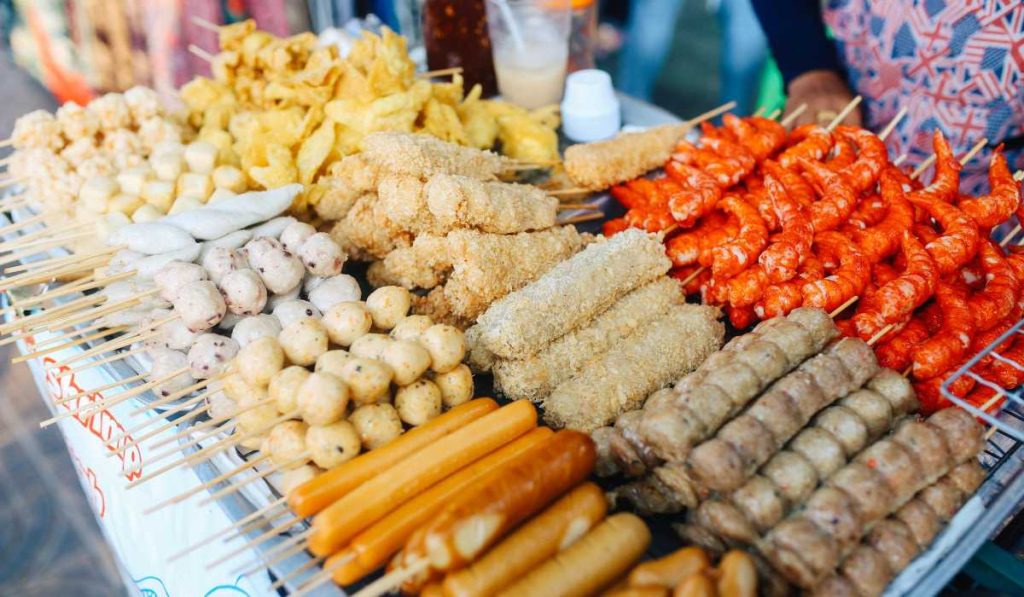 A vibrant display of Bangkok street food with an array of skewered meats, including chicken and shrimp, alongside bowls of noodles, all arranged on a table with a person in the background, epitomizing the city's bustling street food scene.